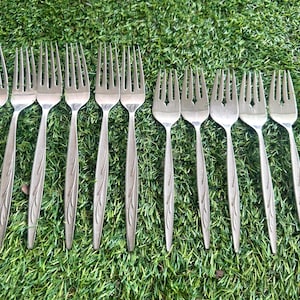 May include: A collection of stainless steel forks with a leaf design on the handles. The forks are arranged on a bed of green grass. The tines of the forks are visible.