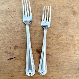 May include: Two silver-toned metal forks of different sizes, each featuring a four-tine head and a handle decorated with vertical fluted lines and a small, ornate scroll-like motif at the base of the handle, showcasing a polished finish.