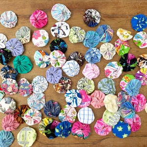 Könnte beinhalten: Eine Sammlung bunter Stoff-Yo-Yos in verschiedenen Größen und Mustern, darunter Blumen- und geometrische Designs. Die Yo-Yos sind auf einer Holzoberfläche angeordnet und zeigen eine Vielzahl von Farben wie Rosa, Blau, Grün und Weiß.
