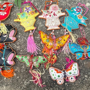 Pode incluir: Uma coleção de ornamentos bordados coloridos em várias formas, incluindo pássaros, borboletas e desenhos florais. Cada ornamento apresenta costuras intrincadas, detalhes de contas e borlas decorativas. Os ornamentos são feitos de tecido e provavelmente feitos à mão.