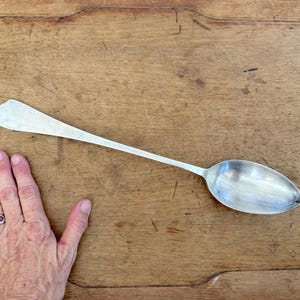 Könnte beinhalten: Ein langstieliger Löffel aus silberfarbenem Metall mit einer glatten, ovalen Laffe und einem flachen, leicht ausgestellten Griffende. Der Löffel hat eine polierte Oberfläche und ein schlichtes, schmuckloses Design über die gesamte Länge.