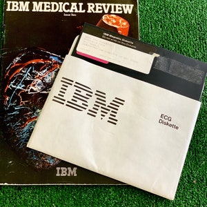 May include: A white envelope with the IBM logo printed in black. The envelope contains a black floppy disk with a white label that reads "IBM Magnetic Diskette Do not write on this Diskette 5735-14-2924 V-L 010410 DEMONSTRATION DISKETTE-DO NOT ERASE 041982 DATE 04-21-02" and "ECG Diskette". The envelope is sitting on top of a magazine titled "IBM MEDICAL REVIEW Issue Two".
