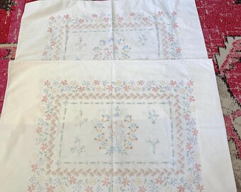 May include: Two white pillowcases with a floral pattern. The design features a rectangular border of pink and light brown flowers and a central arrangement of blue and pink flowers.