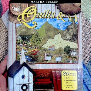 Op de afbeelding: Boekomslag met een gele titel "Small Quilts & Such" van Martha Pullen. De omslag toont een kleurrijke illustratie van een klein stadje met een kerk, huizen en een berg op de achtergrond. Het boek bevat 20 projecten, waaronder quilts, wandkleden, kussens, een placemat en een ovenwant.