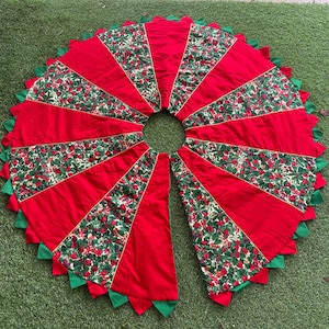 May include: A circular Christmas tree skirt featuring alternating panels of solid red fabric and a festive green and red holly berry print. The edge is finished with a scalloped border of green fabric triangles, and a center opening is provided.