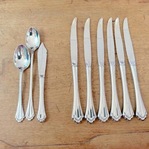 May include: A selection of silver-coloured cutlery, including spoons and knives, arranged on a wooden surface. The spoons have rounded bowls, and the knives have serrated blades. The cutlery is likely for dining.