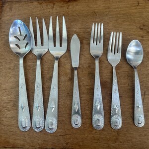 May include: A set of six silver-toned metal utensils including a slotted serving spoon, two large forks, a small butter knife, a medium fork, a small fork, and a small spoon. Each handle features a repeating embossed diamond and oval pattern.