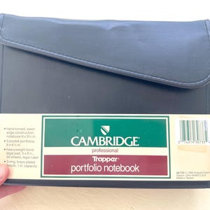 May include: A black Cambridge Professional Trapper portfolio notebook with a flap closure. The packaging label lists features including hand-turned sewn edge construction, 3 pocket portfolios, a 50-sheet legal ruled 6 by 8 inch pad, and a 1 inch 3-ring fixture.
