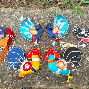 Pode incluir: Uma coleção de ornamentos de galo coloridos e feitos à mão. Os ornamentos apresentam cores vibrantes como vermelho, azul, amarelo e laranja, com detalhes intrincados e costuras decorativas. Cada ornamento tem uma corda para pendurar.