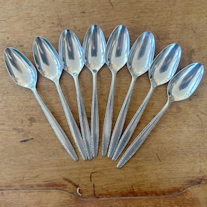 May include: A collection of eight silver spoons arranged in a fan shape on a wooden surface. The spoons have a classic design with elongated bowls and detailed handles. The spoons are reflective, and the wood has a warm, natural tone.