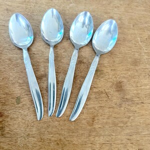 May include: A set of four silver-toned metal spoons with a polished finish. Each spoon features a smooth, curved handle that tapers toward the end and a subtle, embossed floral pattern located at the base of the bowl where it meets the handle.