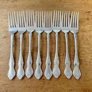 May include: A collection of nine silver forks with ornate floral detailing on the handles. The forks are arranged in a row on a wooden surface, showcasing their elegant design and vintage aesthetic. Ideal for a formal dining setting.