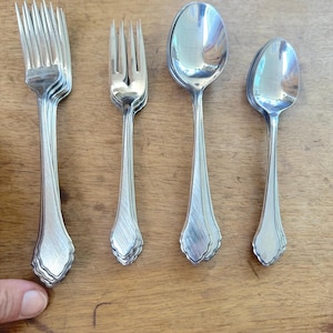 Könnte beinhalten: Eine Sammlung silberfarbener Bestecke, darunter Gabeln und Löffel, die auf einer Holzoberfläche angeordnet sind. Die Gabeln haben mehrere Zinken, und die Löffel haben ovale Schalen. Das Besteck hat ein dekoratives Muster auf den Griffen.