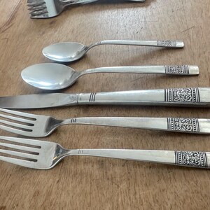 May include: A set of stainless steel flatware including two forks, one dinner knife, and two spoons. Each piece features a smooth handle with a decorative black and silver etched floral pattern near the base and three horizontal lines above it.