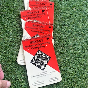 May include: A stack of red and white spiral-bound notepads with the text "BRYANT Machinery & Engineering Company" printed on the cover. The notepads are on a bed of green grass. The notepads also include the address and phone number.
