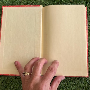May include: An open book with blank, cream-colored pages. The book has a red cover with a gold floral pattern. A hand with a gold ring is holding the book open on a green surface.