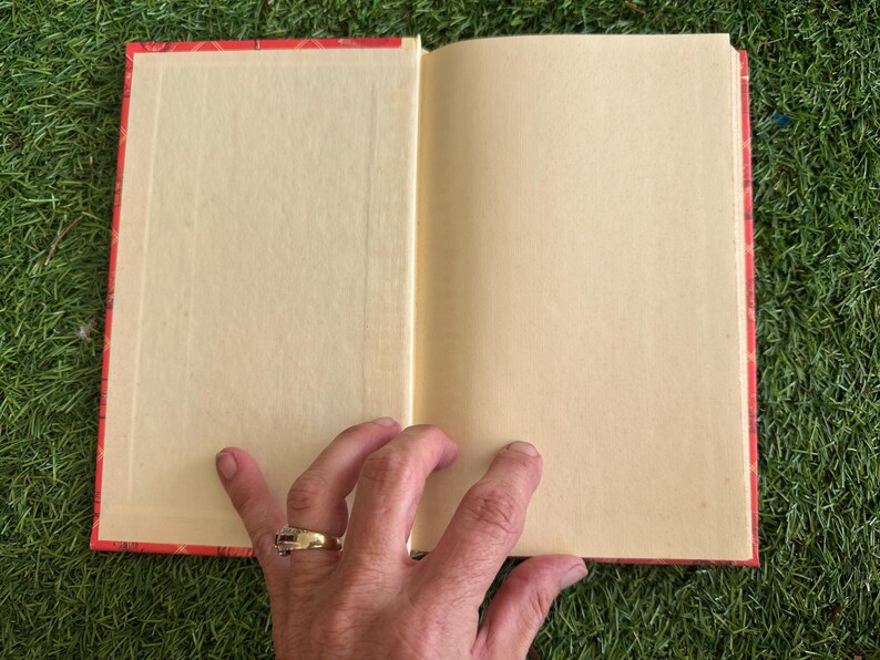 May include: An open book with blank, cream-colored pages. The book has a red cover with a gold floral pattern. A hand with a gold ring is holding the book open on a green surface.