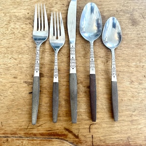 May include: A set of five vintage silverware pieces, including forks, a knife, and spoons. The silverware features ornate detailing on the handles and a mix of silver and dark brown tones. The utensils are arranged on a wooden surface.