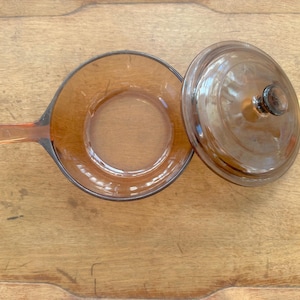May include: Amber-coloured glass saucepan with a handle and a matching lid. The pan has a circular base and a dark rim. The lid has a knob on top. The pan and lid are on a wooden surface.