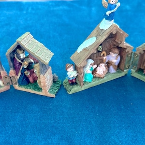 May include: A collection of miniature Christmas nativity scenes. Each scene features a small wooden structure with figures representing the biblical story. The figures are painted in various colors, and the scenes are set on a blue surface.