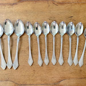 May include: A collection of eleven silver spoons arranged on a wooden surface. The spoons have oval bowls and decorative handles with a fan-like design. The spoons are reflective, and the wood grain is visible.