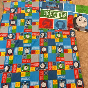 May include: A children's bed sheet set featuring Thomas the Tank Engine and friends. The sheets are patterned with blue, red, and green squares, with images of the trains and the words "PEEP", "STEAM", "SODOR", and "TRUE BLUE".