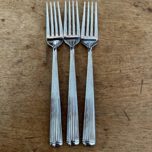 May include: Three silver-colored metal forks arranged side-by-side. Each fork features a polished finish with four tines and a handle decorated with vertical fluted lines that extend from the neck down to a smooth, rounded base at the bottom.