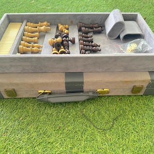 May include: A chess set with a gray velvet-lined storage case. The case holds light and dark chess pieces, dice, and a felt-covered tube. The case is tan and gray with gold-colored clasps and a handle.