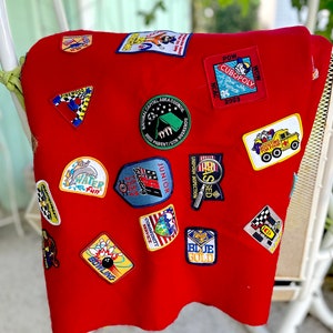 Op de afbeelding: Rode fleece deken met verschillende geborduurde patches, waaronder "Pinewood Derby", "Water Fun", "Junior", "Community Service", "Bowling", "Blue Gold", "POW WOW Cubopoly NCAC 2003" en "Shooting for Food".