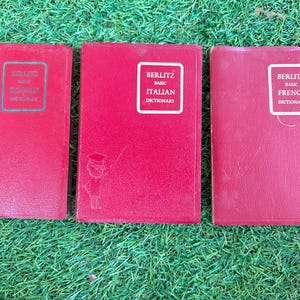 May include: Three vintage Berlitz Basic Dictionary books in red covers. The books are for German, Italian, and French. Each book has a white label with the Berlitz logo and the language name. The books are on a green surface.