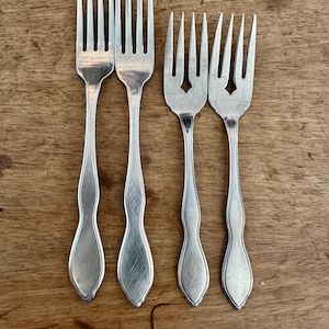 May include: Four silver forks with tines and decorative handles. Two forks have a classic design, while the other two feature a diamond-shaped cutout on the handle. The forks are arranged on a wooden surface.