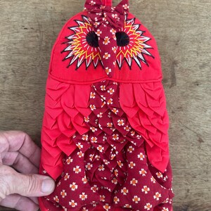 May include: A fabric oven mitt shaped like an owl in shades of red and maroon. The owl features layered fabric feathers, embroidered eyes with yellow and black stitching, and a small fabric bow at the top. The body has a floral pattern.