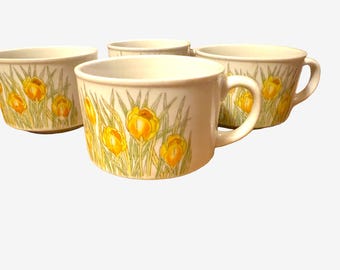 May include: A set of four off-white ceramic mugs featuring a repeating botanical illustration of yellow crocus flowers with green stems and leaves. Each mug has a wide, circular opening, a short handle, and a smooth, rounded base.