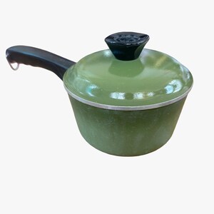 May include: A small olive green saucepan with a matching lid and a black handle. The pot features a silver-toned rim around the top edge, and the lid is topped with a black knob. The handle includes a small metal loop at the end for hanging.