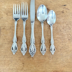May include: A five-piece silver-toned flatware set consisting of two forks, one dinner knife, one large spoon, and one small spoon. Each piece features a polished finish and an ornate, embossed floral pattern along the handle stems.