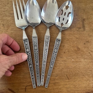 May include: A set of four silver-colored serving utensils: a fork, two spoons, and a slotted spoon. Each handle features a floral design. The utensils are arranged on a wooden surface.