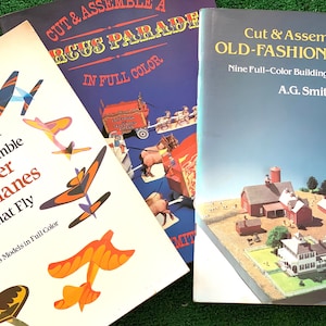 May include: Three papercraft books with colourful covers. The first book is titled "Cut & Assemble Paper Airplanes That Fly" and features a variety of paper aeroplanes. The second book is titled "Cut & Assemble a Circus Parade in Full Colour" and features a colourful circus parade scene. The third book is titled "Cut & Assemble an Old-Fashioned Farm" and features a miniature farm scene with buildings and animals.