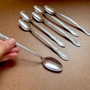 May include: Six silver spoons with ornate handles and a seventh spoon held in a hand. The spoons are arranged in a row on a brown surface.