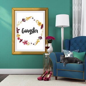 May include: A rectangular gold-toned frame containing a white print with a watercolor floral wreath in shades of yellow, burgundy, and brown, surrounding the word Gangster written in a black, brush-style script font centered on the page.