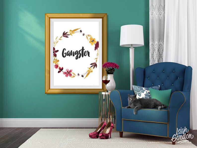 May include: A rectangular gold-toned frame containing a white print with a watercolor floral wreath in shades of yellow, burgundy, and brown, surrounding the word Gangster written in a black, brush-style script font centered on the page.