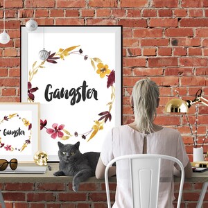 May include: Two framed wall art prints featuring a watercolor floral wreath in shades of yellow, burgundy, and brown surrounding the word Gangster in black script. The prints are displayed on a desk alongside a gray cat and a gold desk lamp.