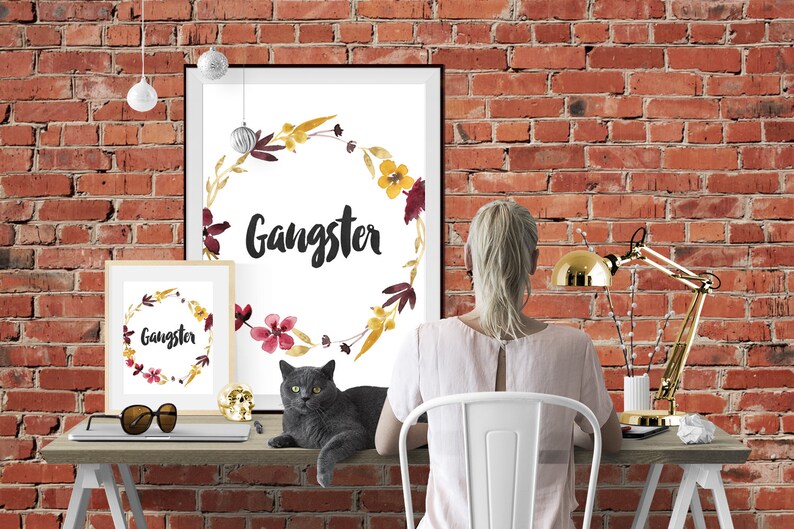 May include: Two framed wall art prints featuring a watercolor floral wreath in shades of yellow, burgundy, and brown surrounding the word Gangster in black script. The prints are displayed on a desk alongside a gray cat and a gold desk lamp.