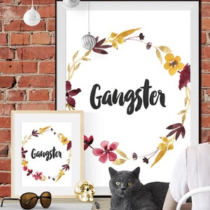May include: Two framed wall art prints featuring the word Gangster in black script, surrounded by a watercolor floral wreath in shades of yellow, burgundy, and brown. The prints are displayed on a desk with a gray cat and gold skull.