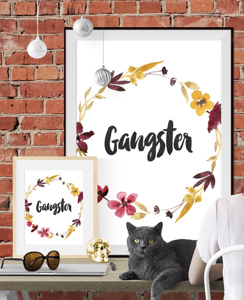 May include: Two framed wall art prints featuring the word Gangster in black script, surrounded by a watercolor floral wreath in shades of yellow, burgundy, and brown. The prints are displayed on a desk with a gray cat and gold skull.
