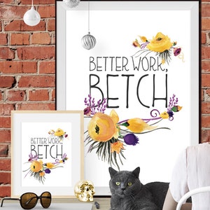 May include: A framed wall art print featuring the text "BETTER WORK, BETCH" in black lettering, surrounded by watercolour illustrations of yellow flowers and purple sprigs. The print is displayed in a black frame, with a smaller matching version nearby.
