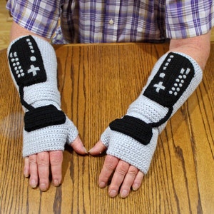 May include: A pair of gray crocheted fingerless gloves with black crocheted game controller designs. The gloves have a cord design on the back of the hand.