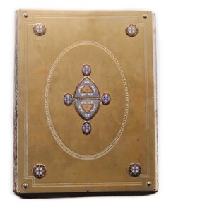 May include: A rectangular, gold-toned book cover with intricate detailing. The cover features an oval design in the center, adorned with colorful geometric patterns. Decorative circular accents are placed at each corner, adding to the ornate aesthetic.