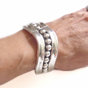 May include: A silver bracelet with a curved design and a row of beads. The bracelet is on a person's wrist. The person is also wearing a silver ring with a textured design.