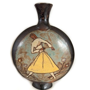 May include: A ceramic vase with a round body and a narrow neck. The vase features a painted design of a person in a yellow dress, with a light blue background and small flowers. The vase has a dark brown rim and base.
