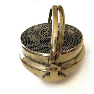 May include: A vintage brass incense burner with a round, perforated top and a hinged handle. The top features decorative cutouts, including a central symbol. The burner has a two-tiered design and a clasp closure.